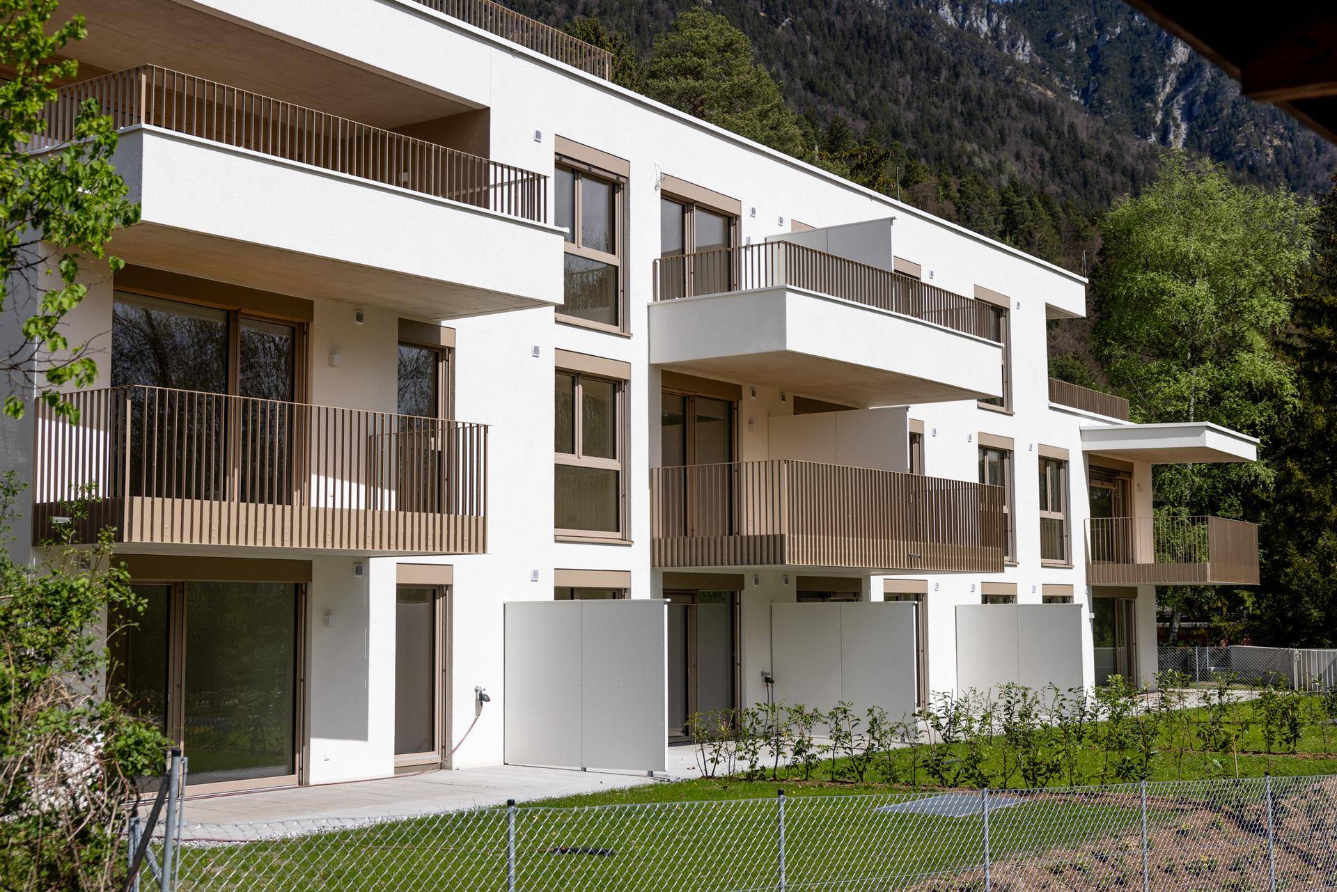 Modernes Mehrfamilienhaus mit mehreren Balkonen und grüner Außenanlage vor Bergkulisse.