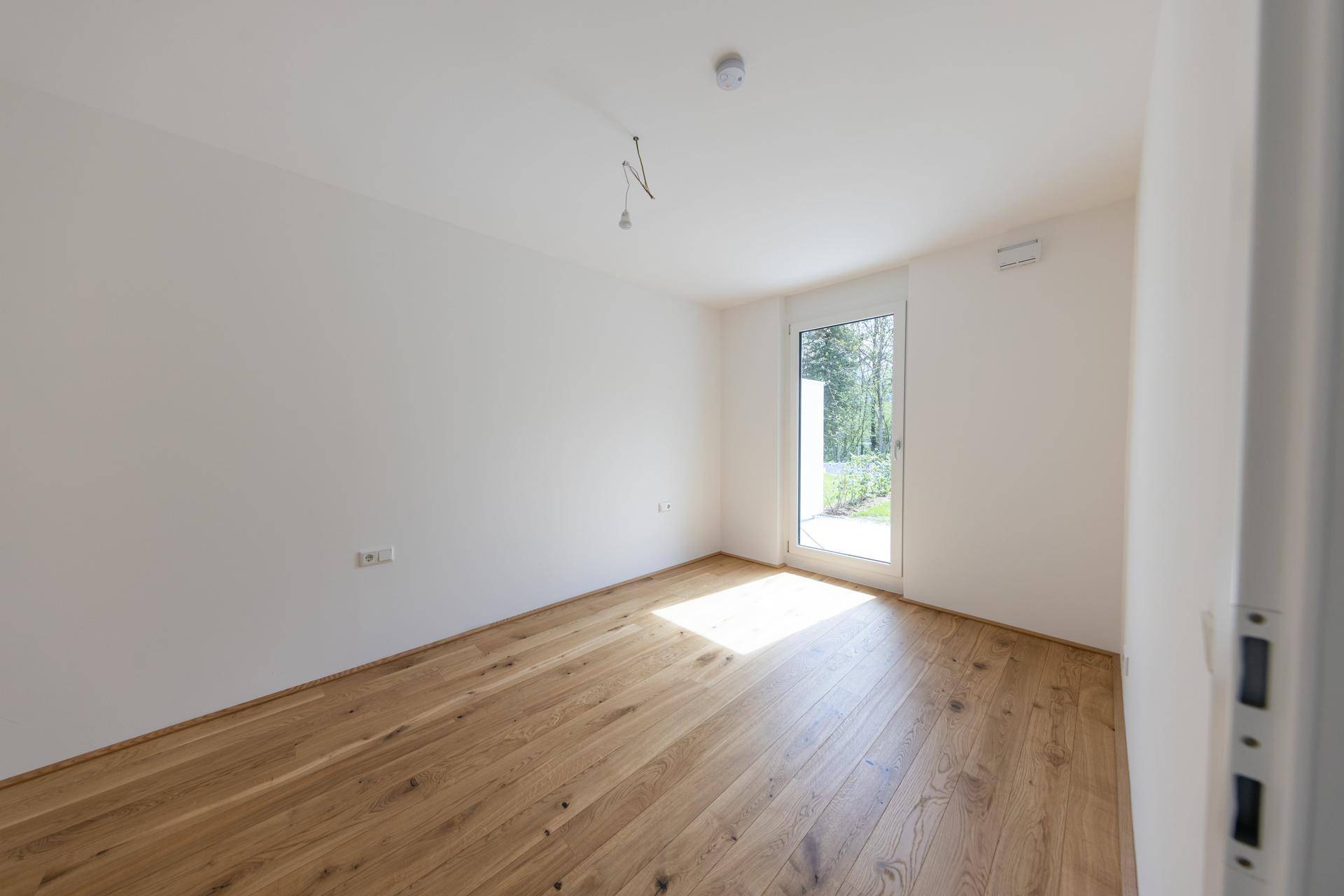 Helles, leeres Zimmer mit Holzboden und Zugang zu einem sonnigen Außenbereich.