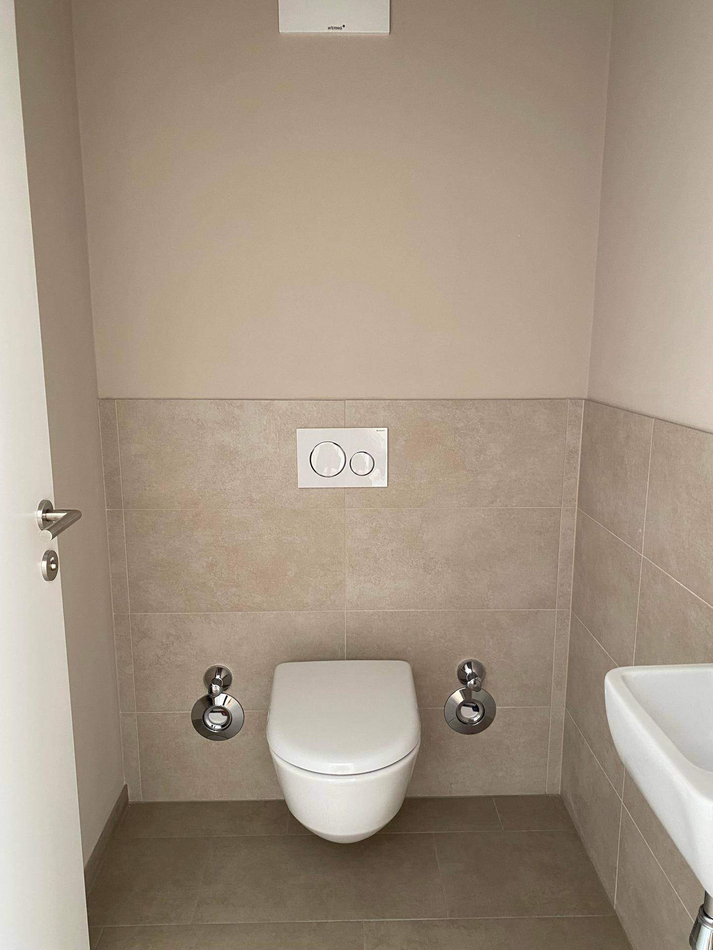 Separate Toilette mit Wand-WC, kleinem Waschbecken und beigefarbenen Fliesen.