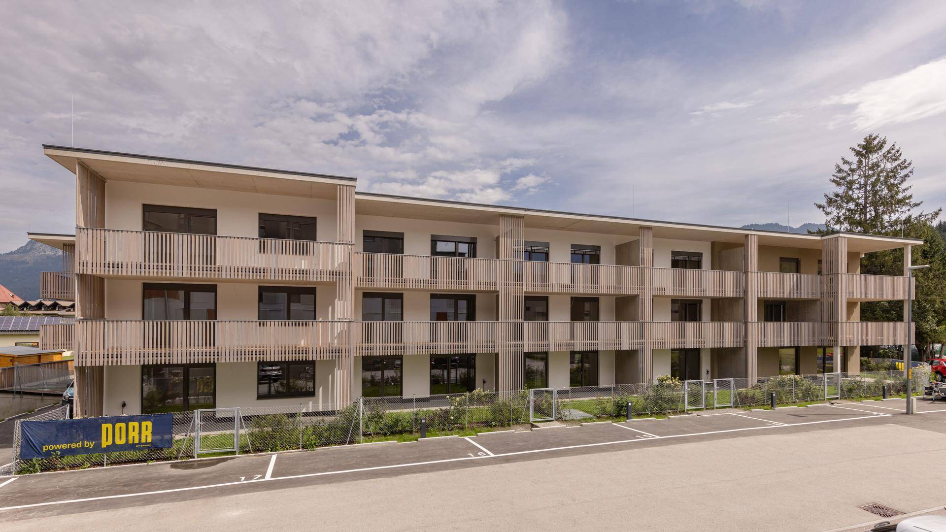 Weitläufiges modernes Wohngebäude mit vielen Balkonen, Holzdetails und Parkplätzen vor Bergkulisse.
