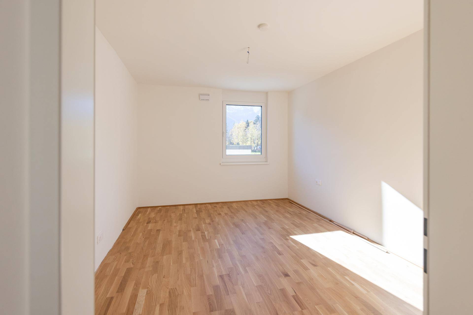 Unmöbliertes Schlafzimmer mit Parkettboden und Fenster, das viel Tageslicht hereinlässt.