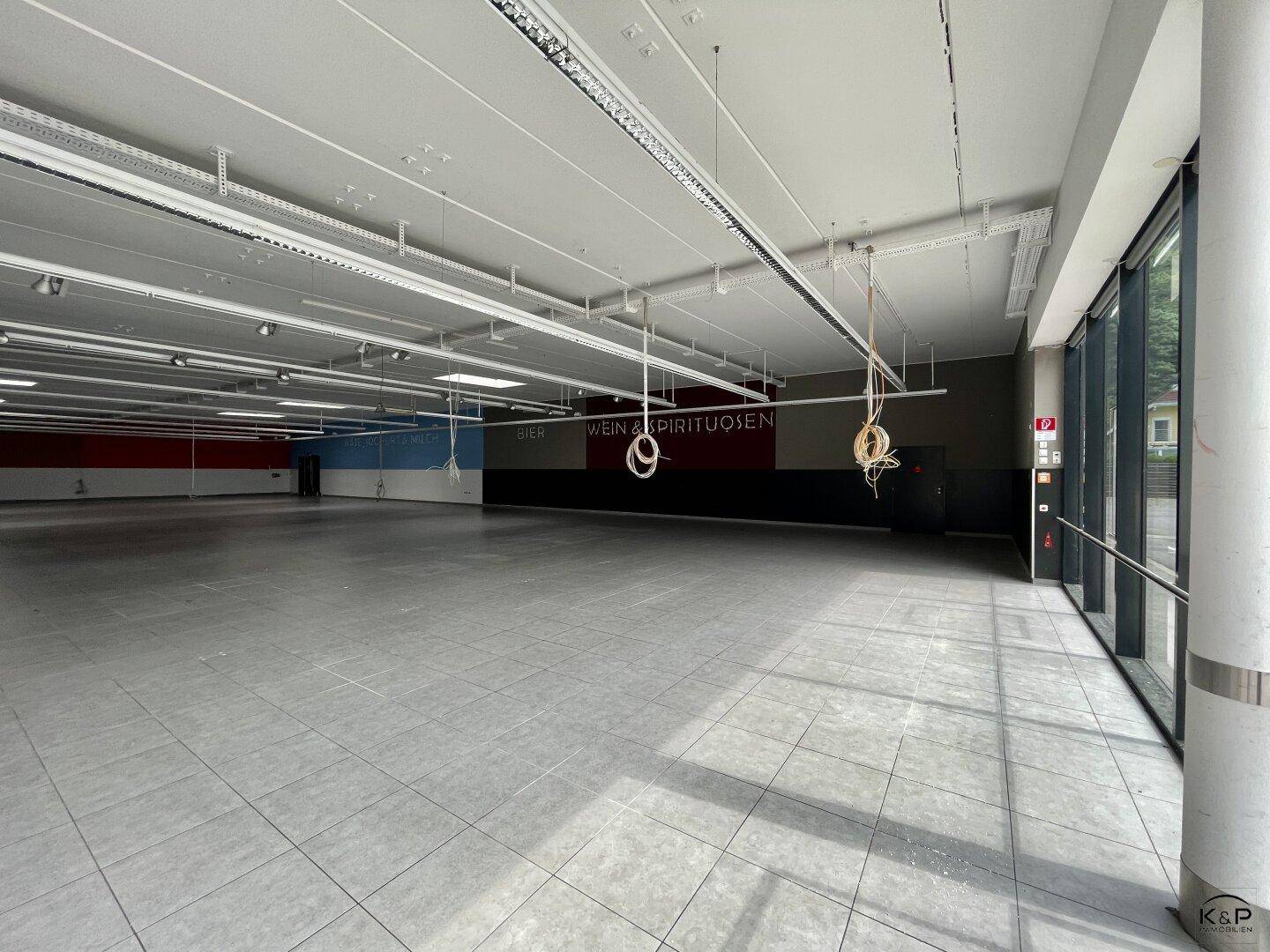 Heller, leerer Ladenraum mit großen Fenstern, Fliesenboden und sichtbaren Deckeninstallationen.