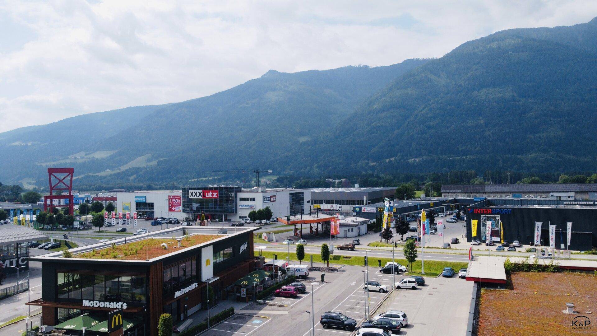 Luftaufnahme eines Gewerbegebiets mit verschiedenen Geschäften und Bergen im Hintergrund.