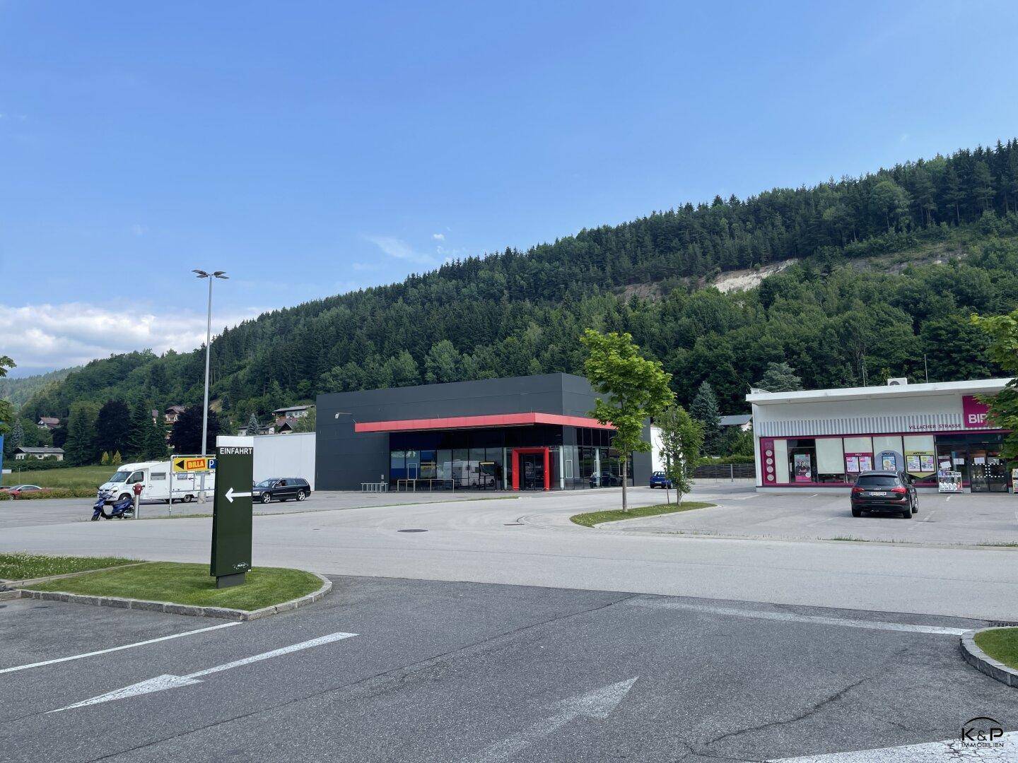 Gesamtansicht des Geschäftsgebäudes mit Parkplatz und umliegender Landschaft mit bewaldeten Hügeln und weiteren Gebäuden.