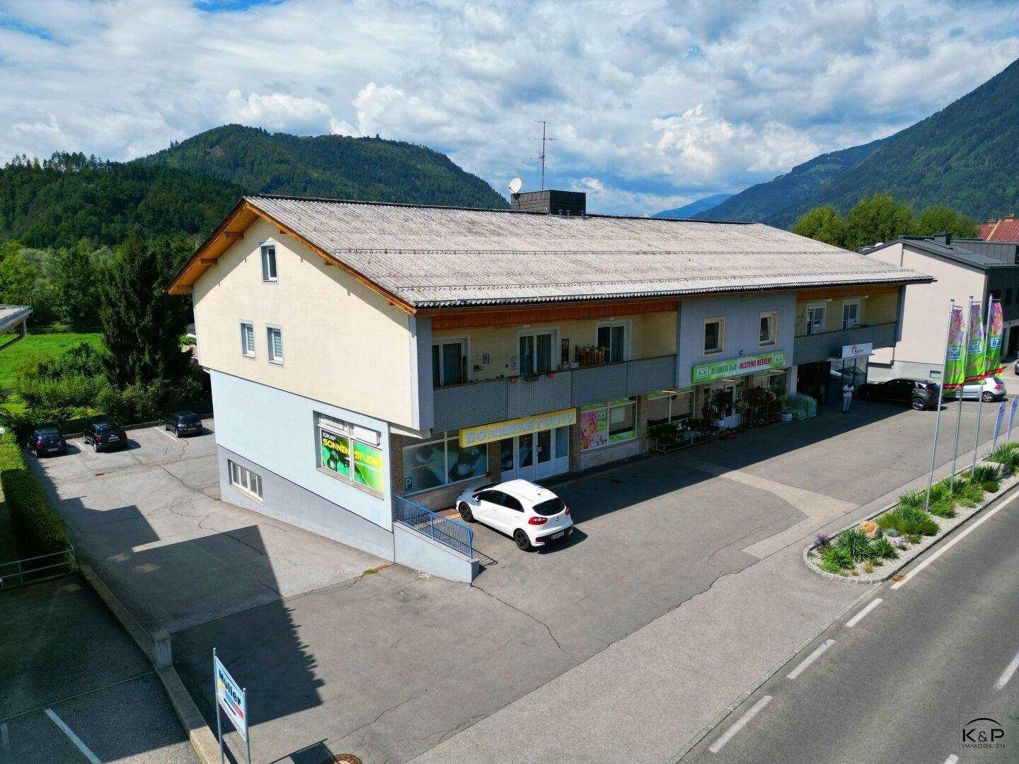 Luftaufnahme des Gebäudes mit Geschäftsflächen im Erdgeschoss und Parkplätzen davor.
