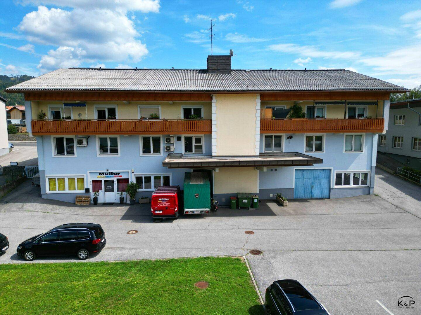 Ansicht der Gebäudefassade mit Balkonen, Geschäften im Erdgeschoss und Parkplätzen davor.