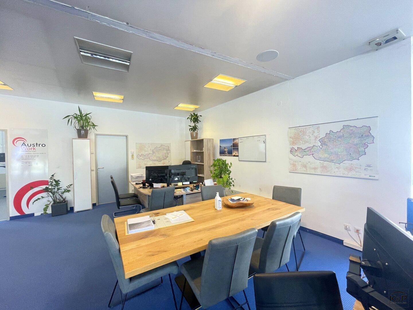 Helles Büro mit großem Holztisch, blauer Teppichboden und Landkarten an den Wänden.