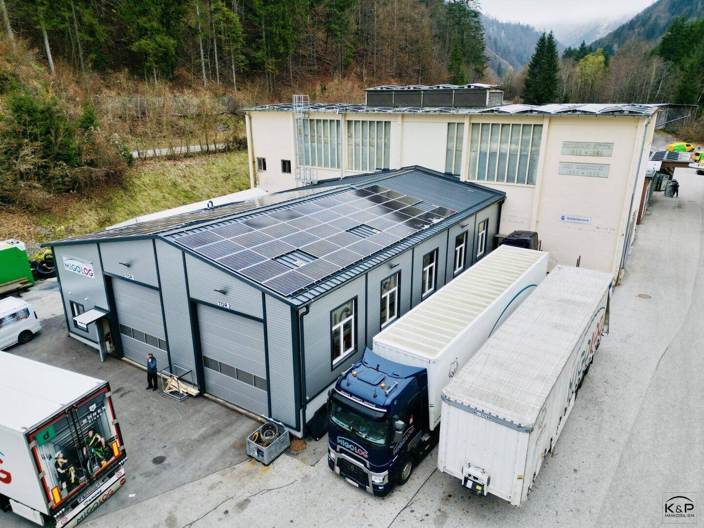 Modernes Industriegebäude mit Rolltoren und Solarpaneelen, umgeben von LKW-Stellplätzen und Wald.