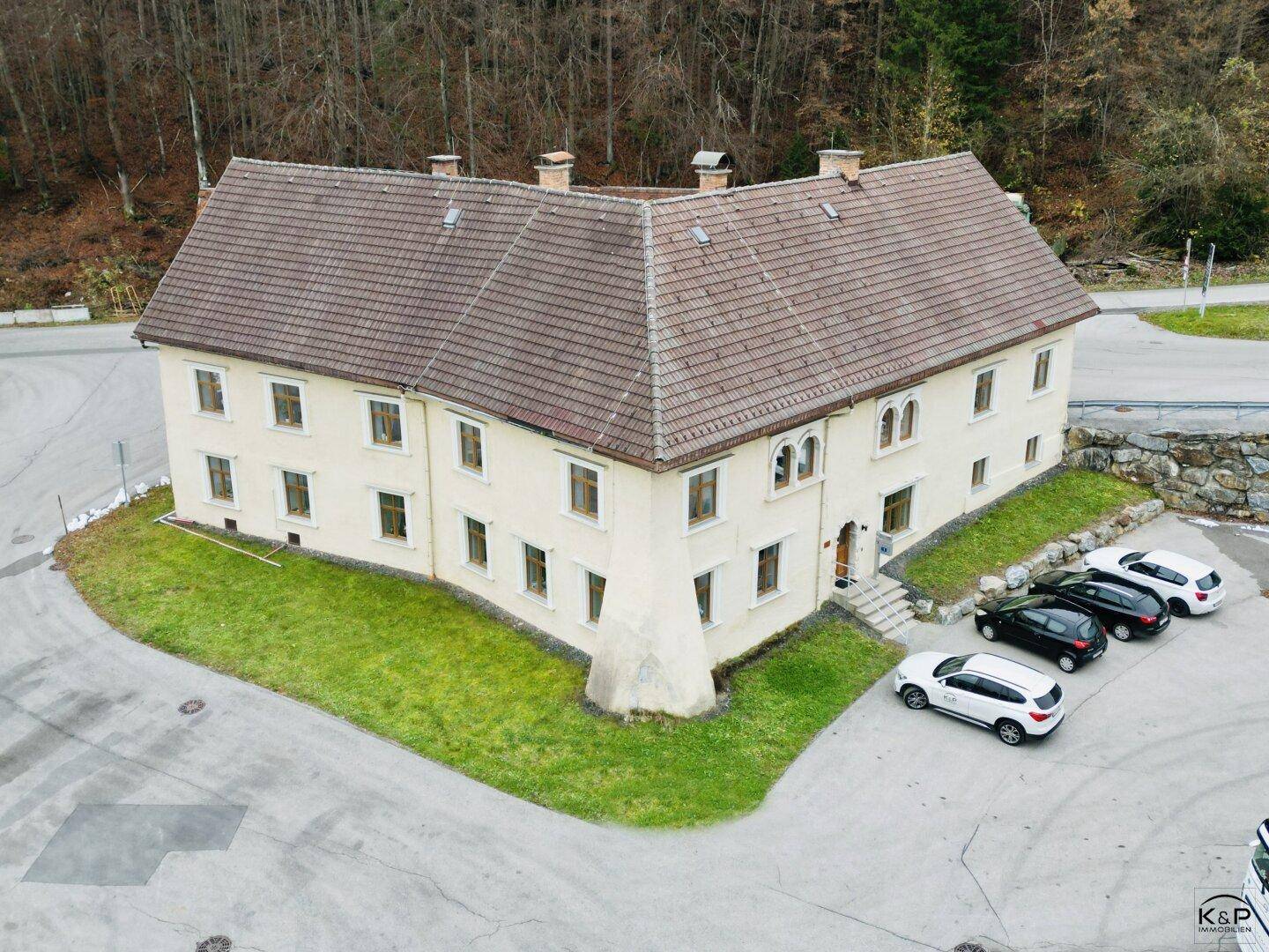 Älteres, traditionelles Gebäude mit heller Fassade und Ziegeldach, umgeben von Grünflächen und Parkplätzen.
