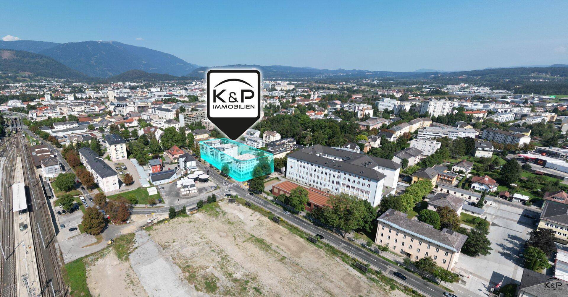 Weitläufige Luftaufnahme der Stadt mit dem markierten Gebäudekomplex und beeindruckendem Bergpanorama im Hintergrund.