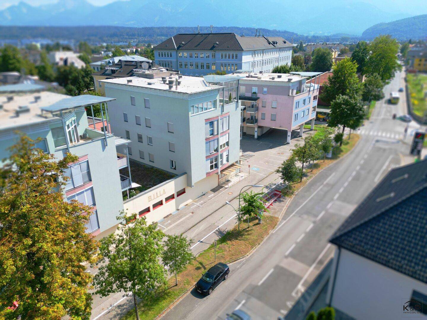 Luftaufnahme des Gebäudekomplexes mit Parkplätzen, umgeben von Bäumen und einer weiten Berglandschaft im Hintergrund.