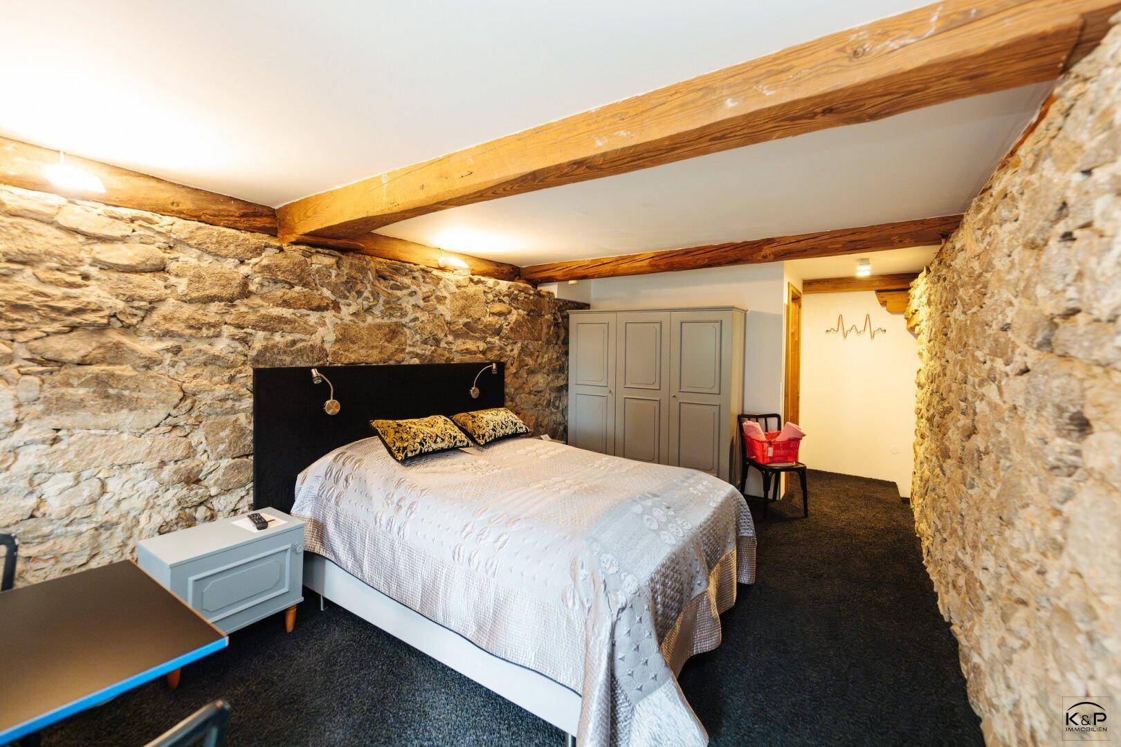 Rustikales Schlafzimmer mit freiliegenden Steinwänden, Holzbalkendecke und dunklem Teppichboden.