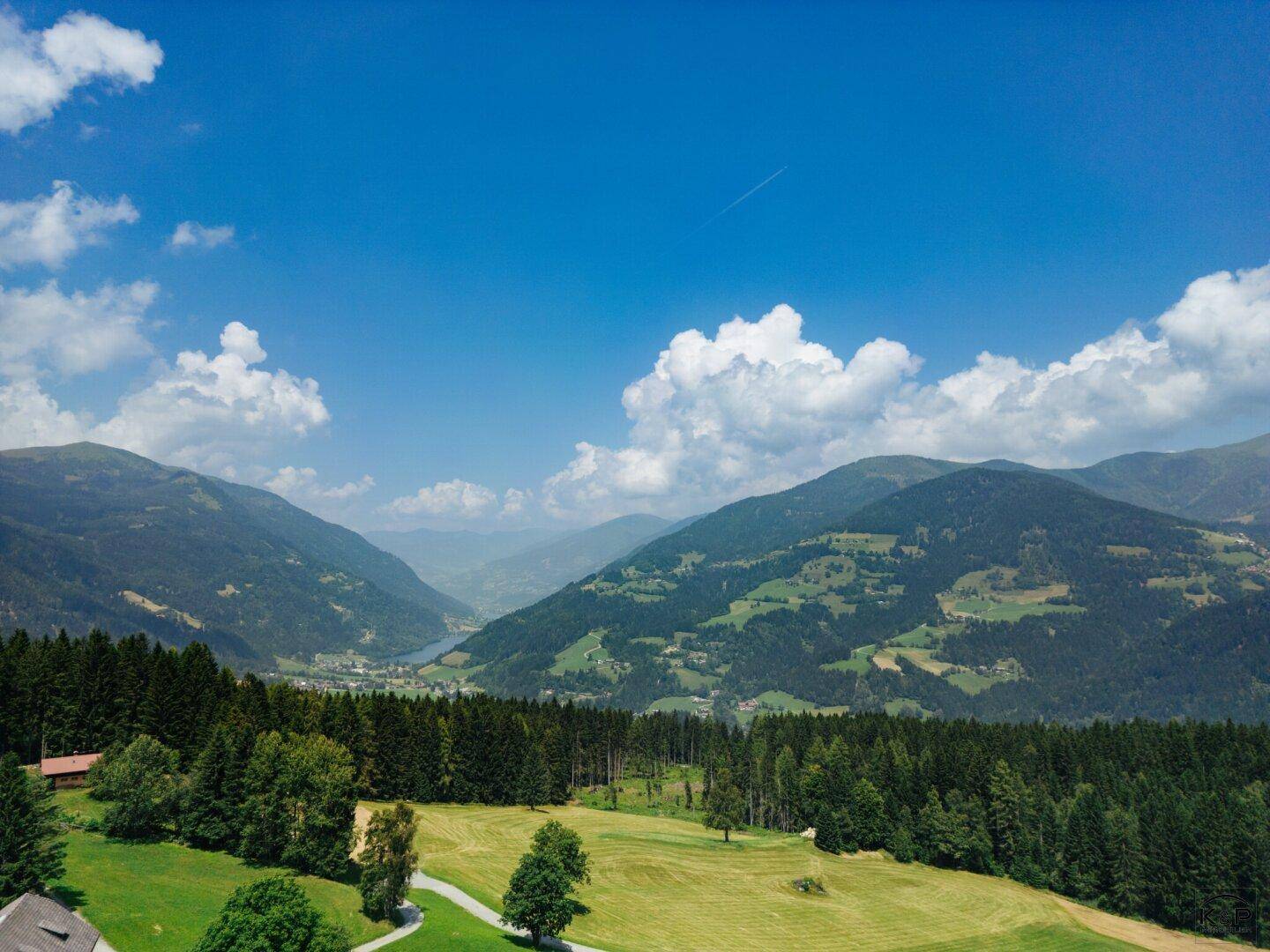 Panoramablick auf eine weite grüne Landschaft mit Wäldern, Bergen und einem See in der Ferne.