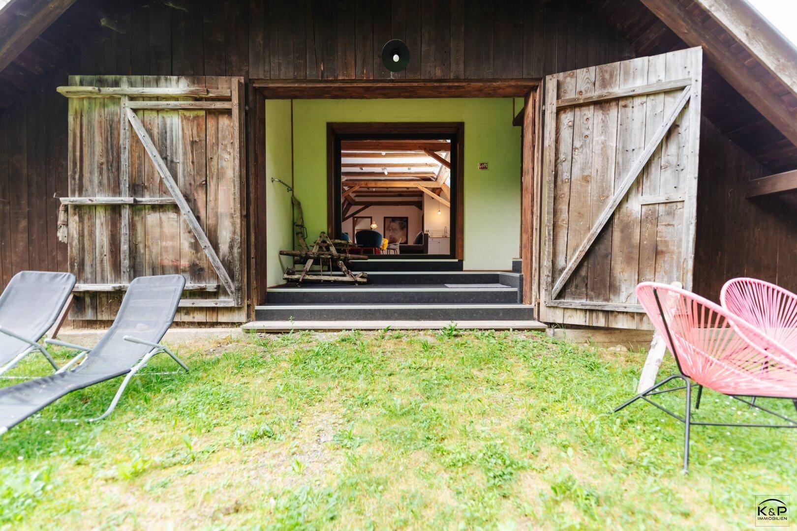 Gartenbereich mit Liegestühlen und Zugang zu einem rustikalen Gebäude mit Holztüren.