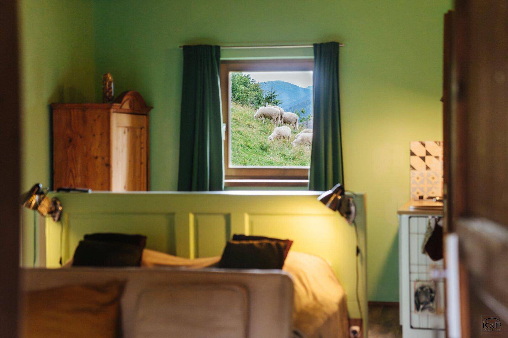 Schlafzimmer mit grünen Wänden und einem Fenster, das den Blick auf weidende Schafe freigibt.