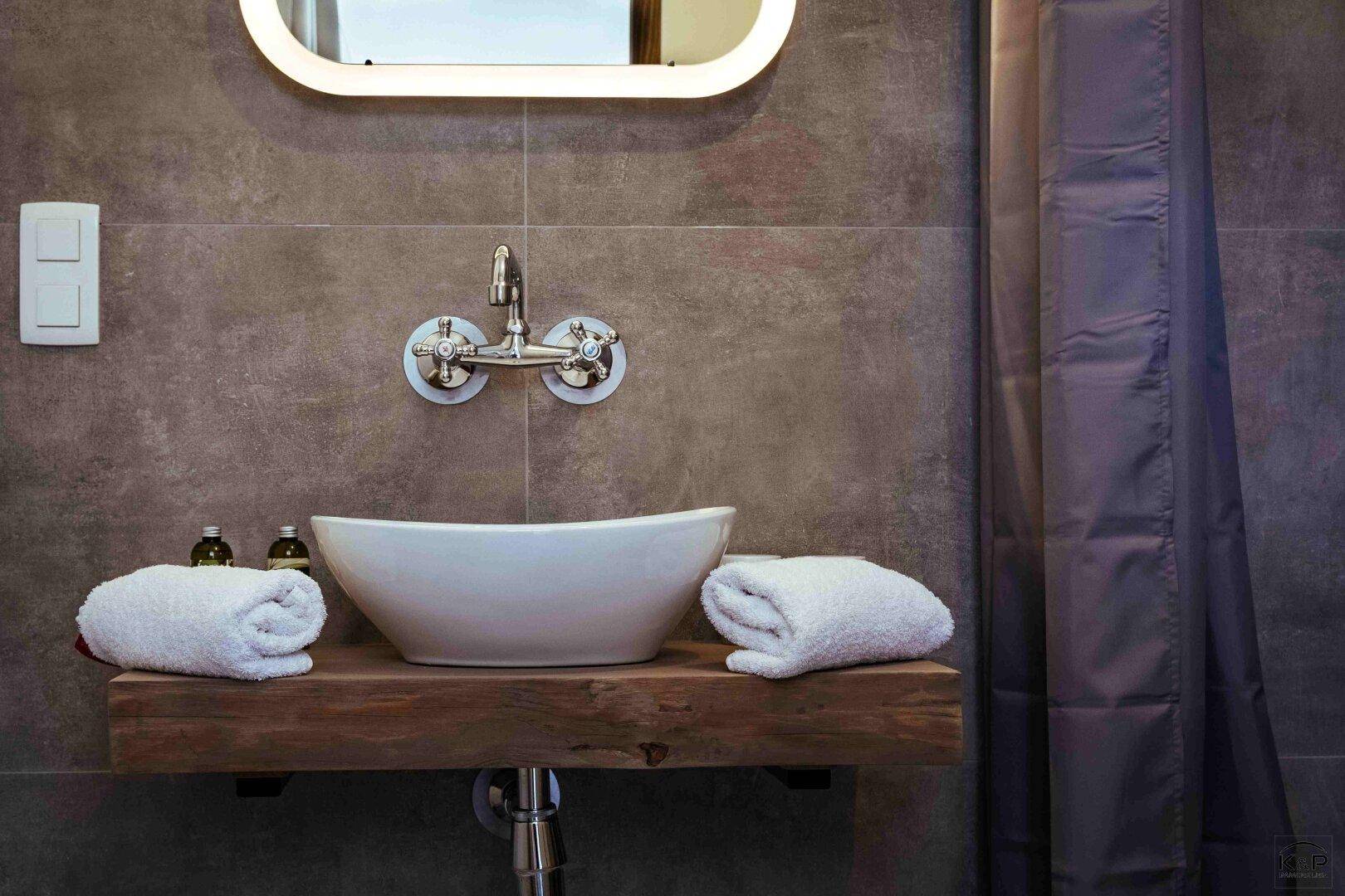 Modernes Badezimmer mit Aufsatzwaschbecken auf Holzplatte und Wandarmatur vor grauen Fliesen.