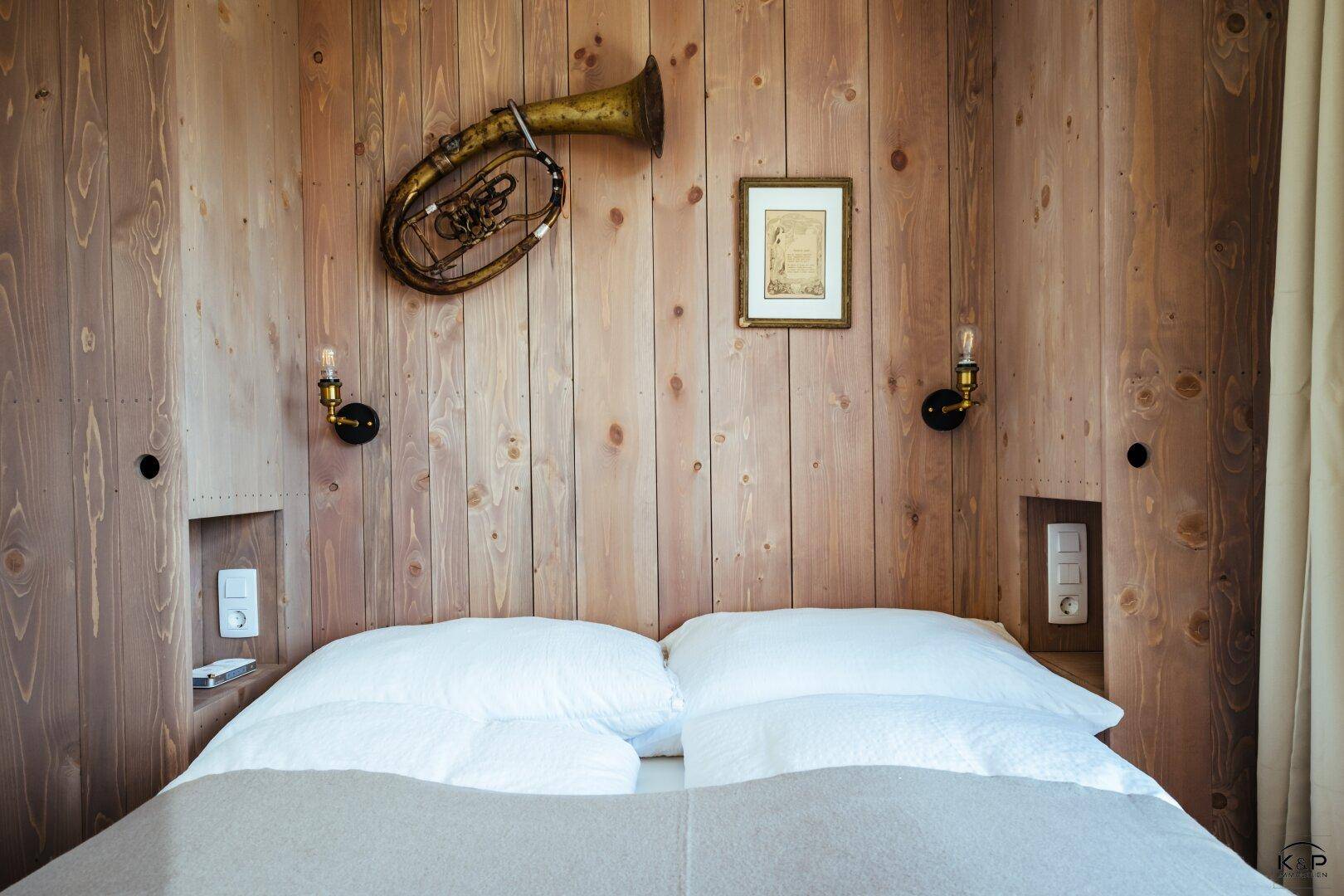 Detailansicht eines Bettes mit Holzkopfteil, Wandleuchten und einem Musikinstrument als Dekoration.