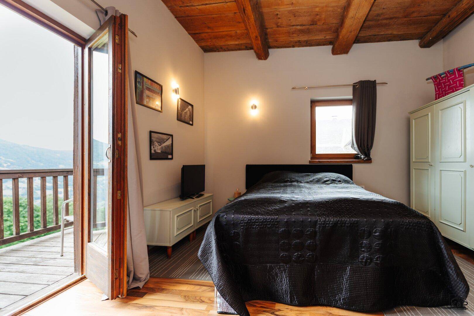 Schlafzimmer mit Holzbalkendecke, Balkonzugang und Blick auf die umliegende Landschaft.