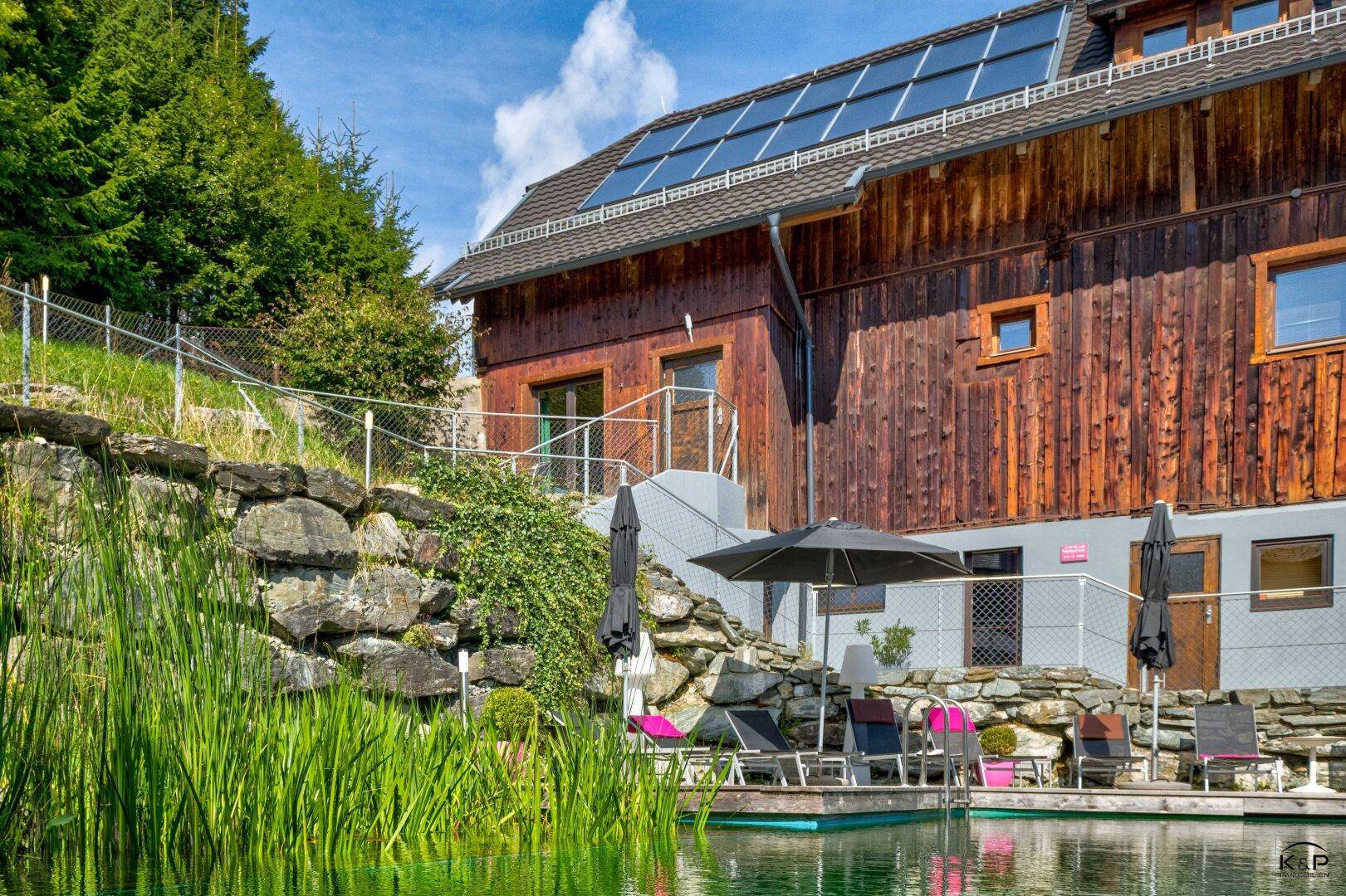 Natürlicher Schwimmteich mit Liegestühlen und einem traditionellen Holzhaus mit Solaranlage.