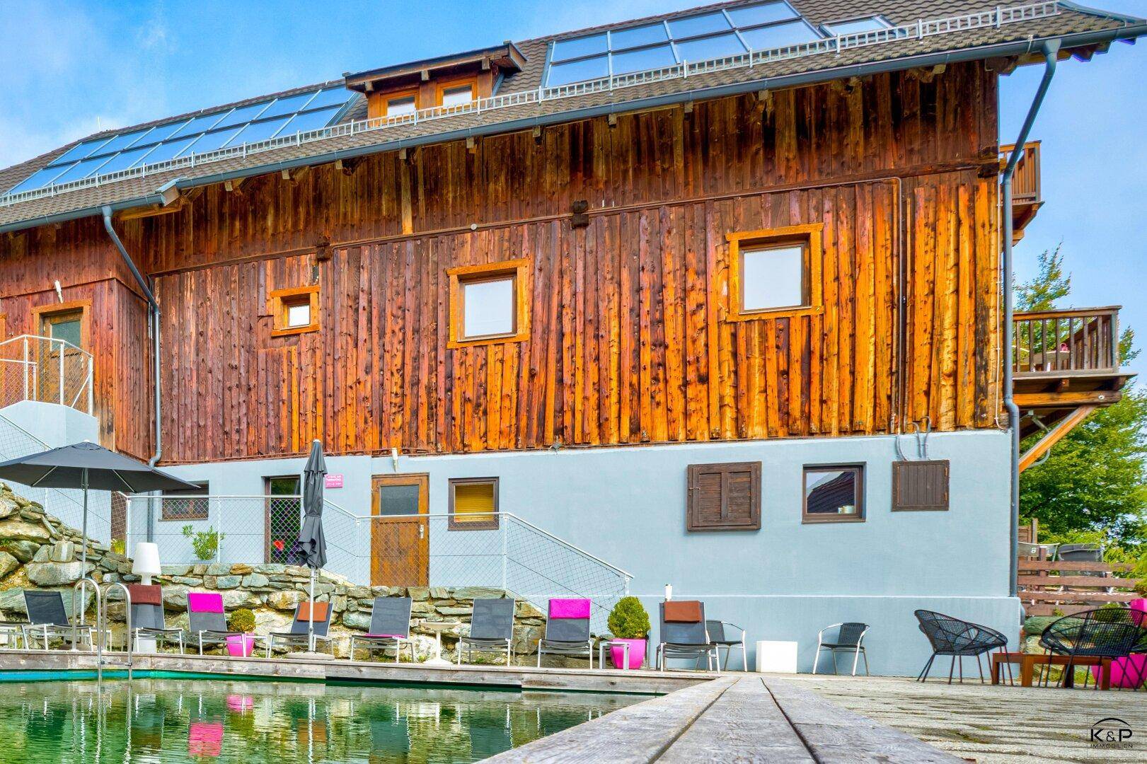 Außenbereich mit Schwimmteich, Liegestühlen und einem Holzhaus mit Solaranlage.