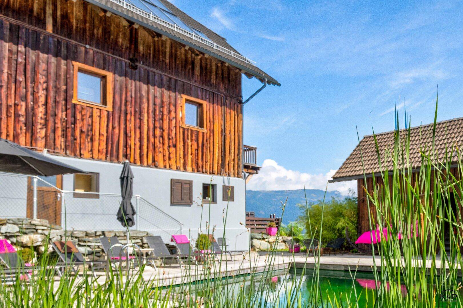 Außenbereich mit Naturpool, Liegestühlen und einem traditionellen Holzhaus im Hintergrund.