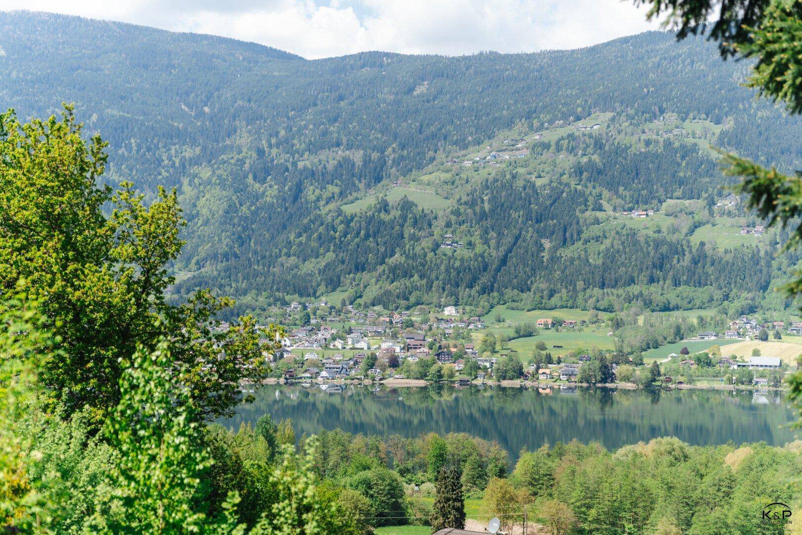 Malerischer Blick auf einen See und ein Dorf, umrahmt von üppigem Grün und Bergen.