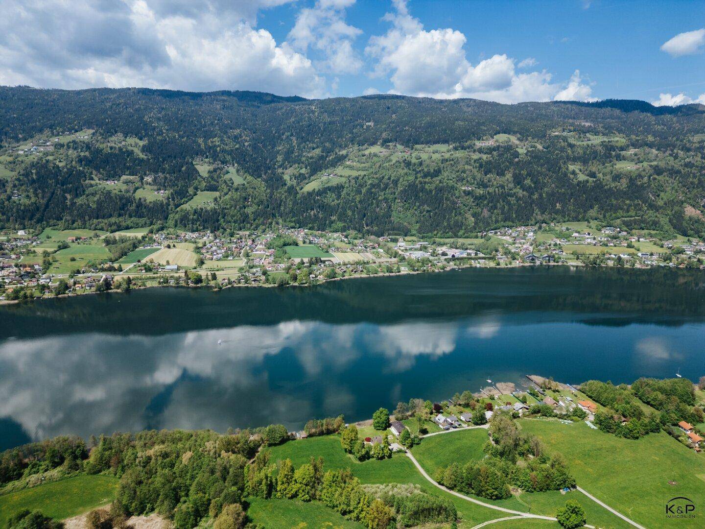 Weitläufige Luftaufnahme eines ruhigen Sees, der von bewaldeten Bergen und einer Siedlung am Ufer gesäumt wird.