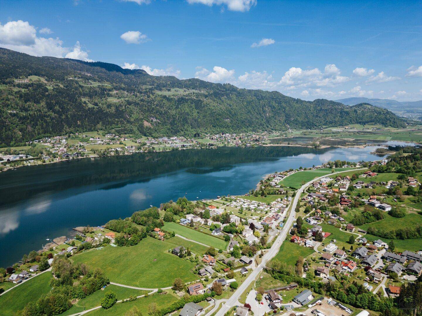 Malerische Luftaufnahme eines Sees mit angrenzender Ortschaft und einer Straße, eingebettet in eine grüne Berglandschaft.