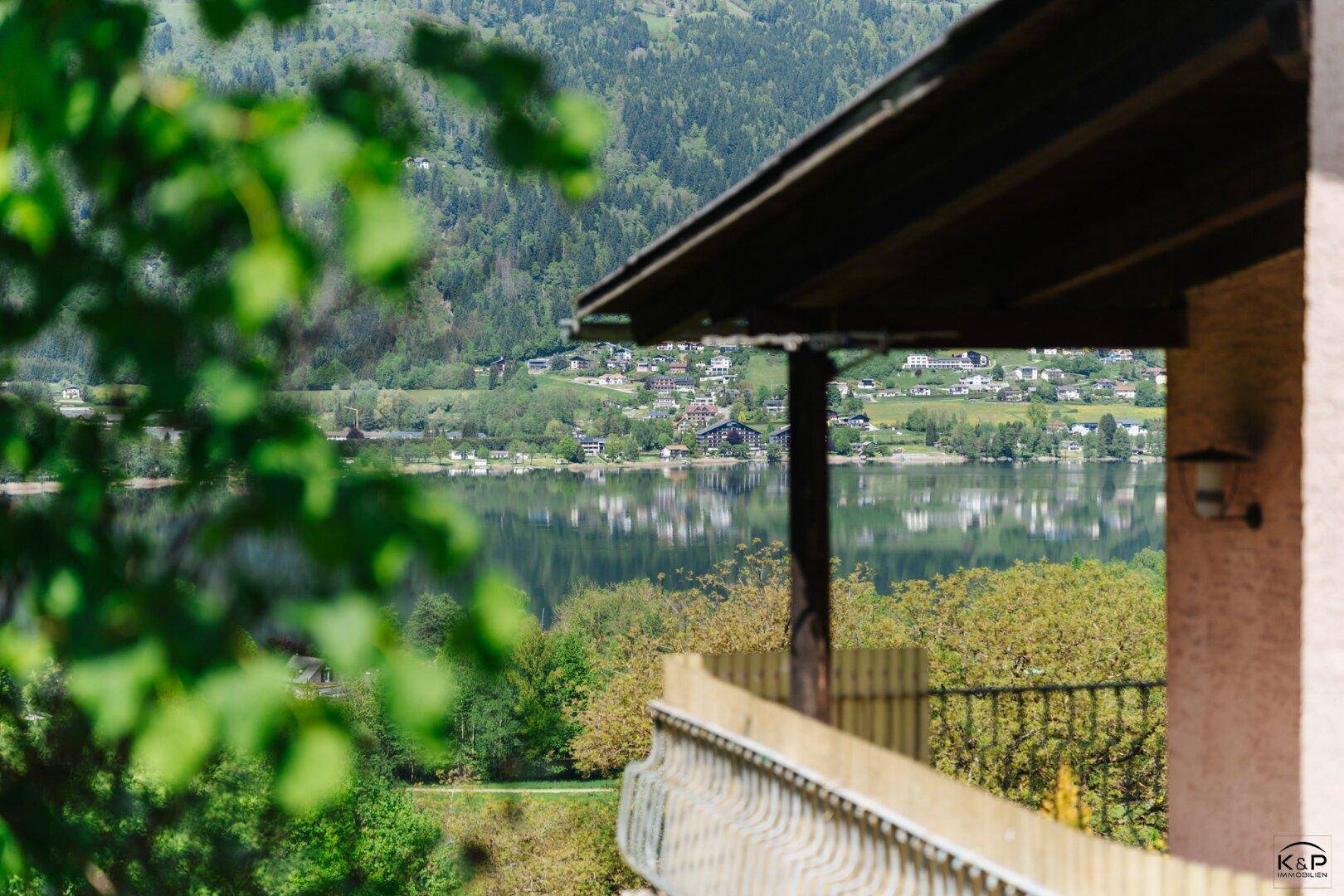Blick von einem Balkon mit Holzgeländer auf einen See und die gegenüberliegende bewaldete Berglandschaft.
