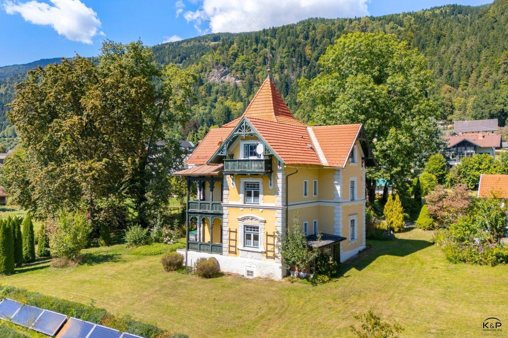Historische Villa mit gelber Fassade, rotem Dach und weitläufigem Garten in alpiner Umgebung.