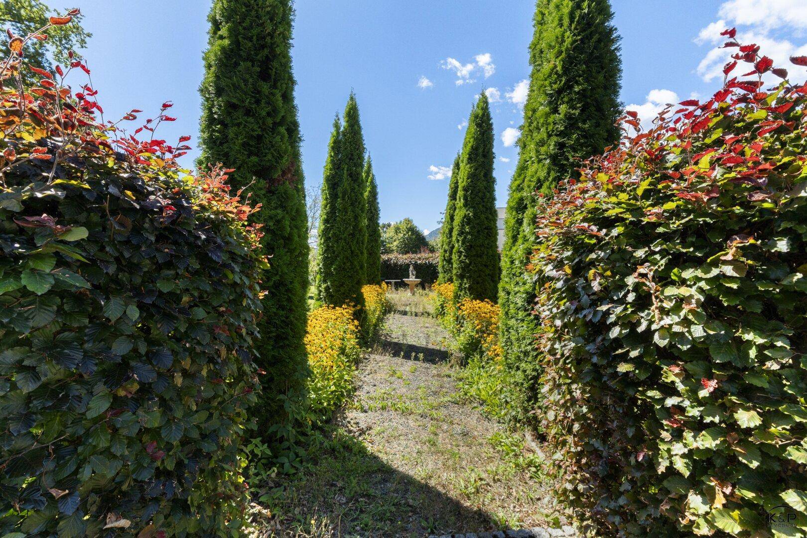 Gepflegter Gartenweg, gesäumt von hohen Hecken und Zypressen, führt zu einem Brunnen.
