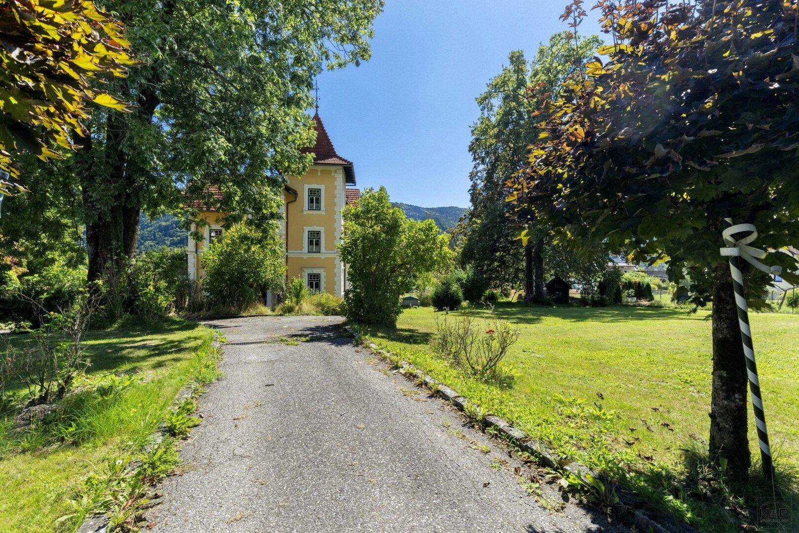 Zufahrt zur Villa, umgeben von altem Baumbestand und weitläufigen Grünflächen.