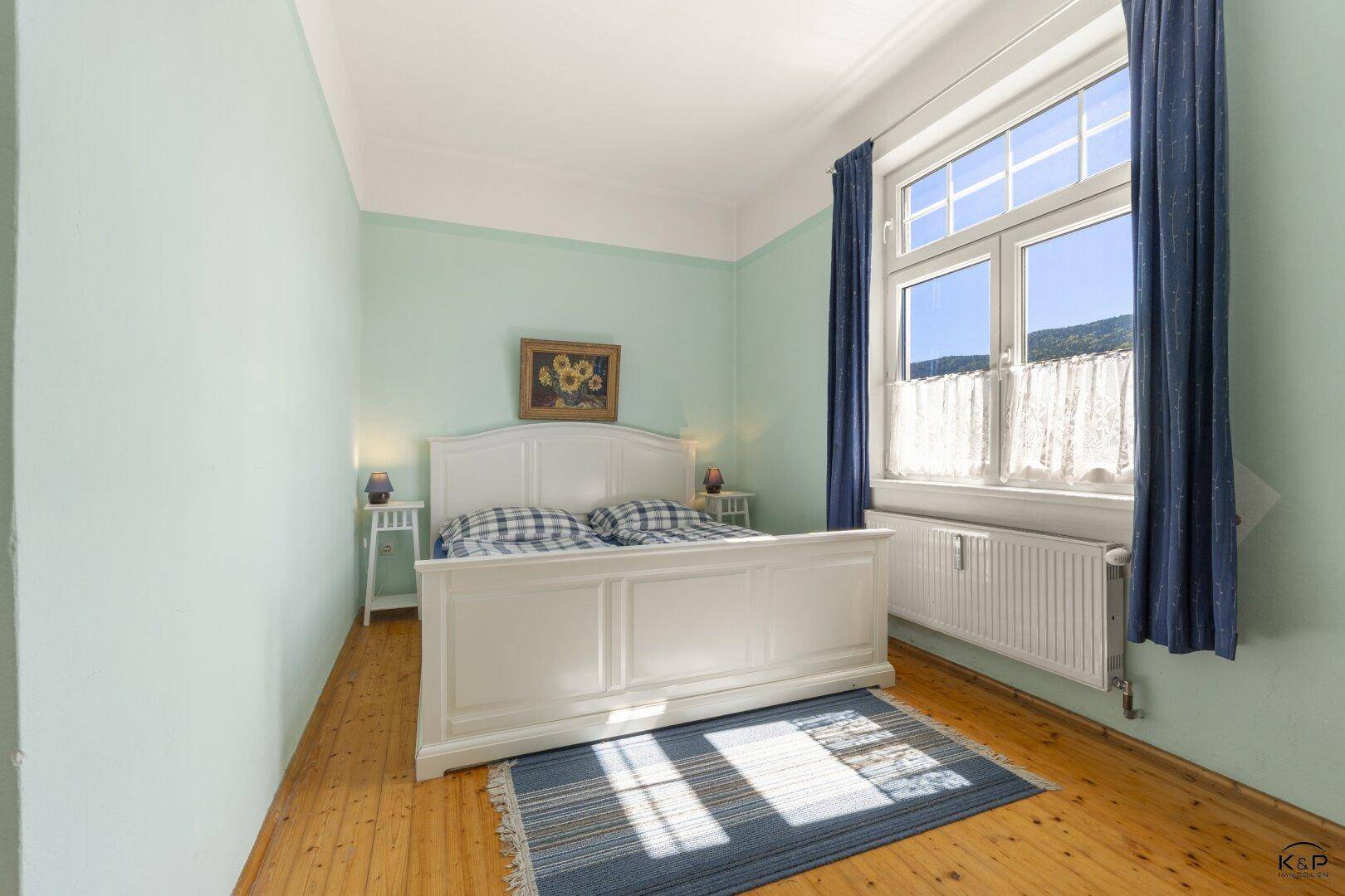 Schlafzimmer mit weißem Bett, hellgrünen Wänden und großem Fenster für viel Tageslicht.