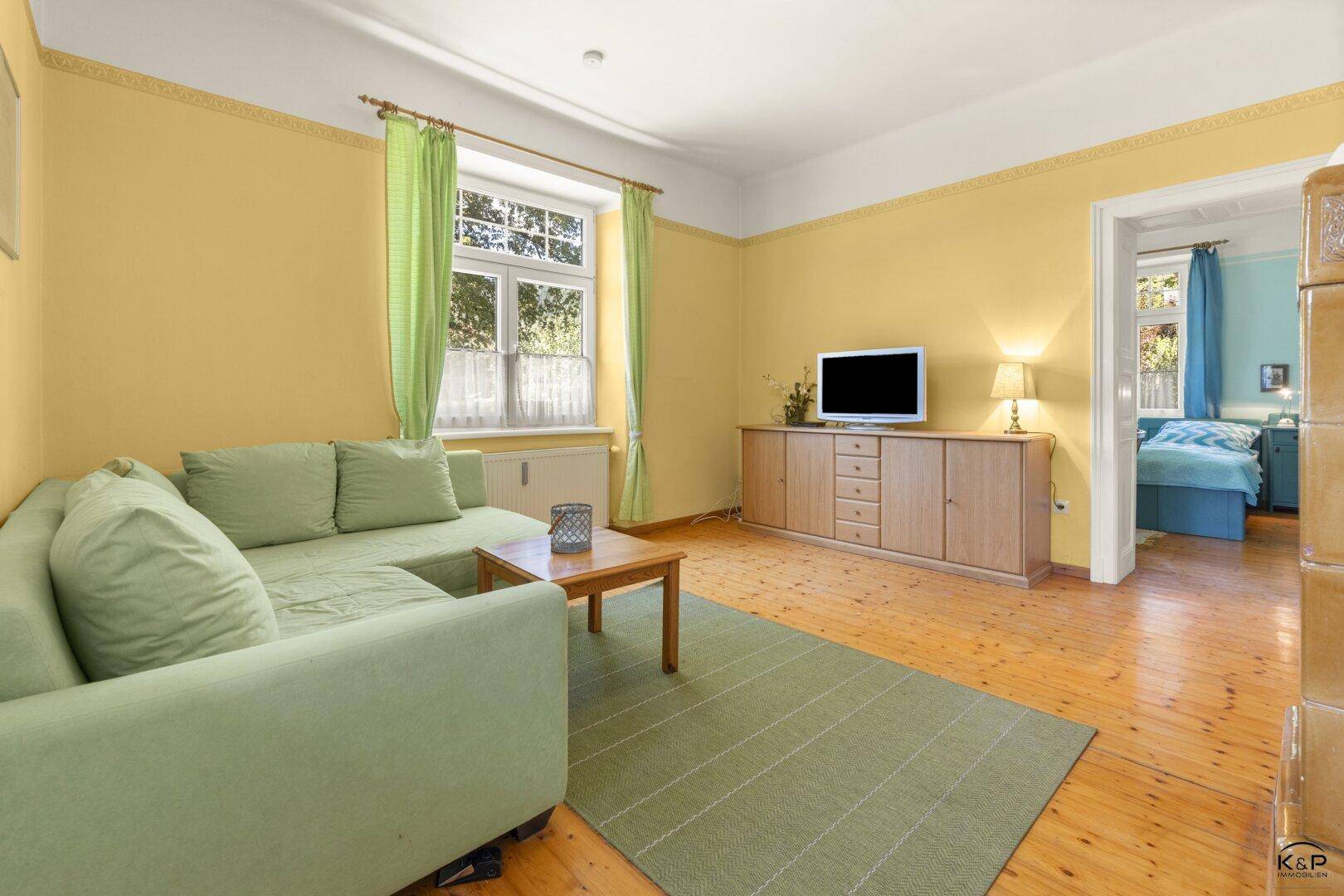Helles Wohnzimmer mit grünem Sofa, Holzboden und Zugang zu einem weiteren Zimmer.