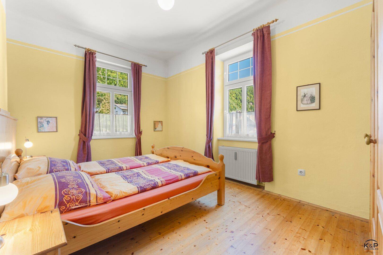 Schlafzimmer mit Holzbett, gelben Wänden und Fenstern mit Blick ins Grüne.