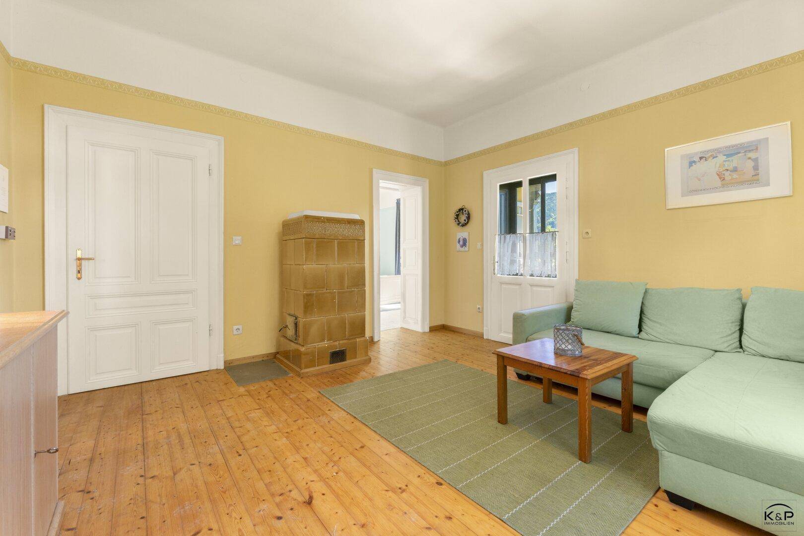 Wohnzimmer mit grünem Sofa, Holzboden und einem hellen Kachelofen als Wärmequelle.