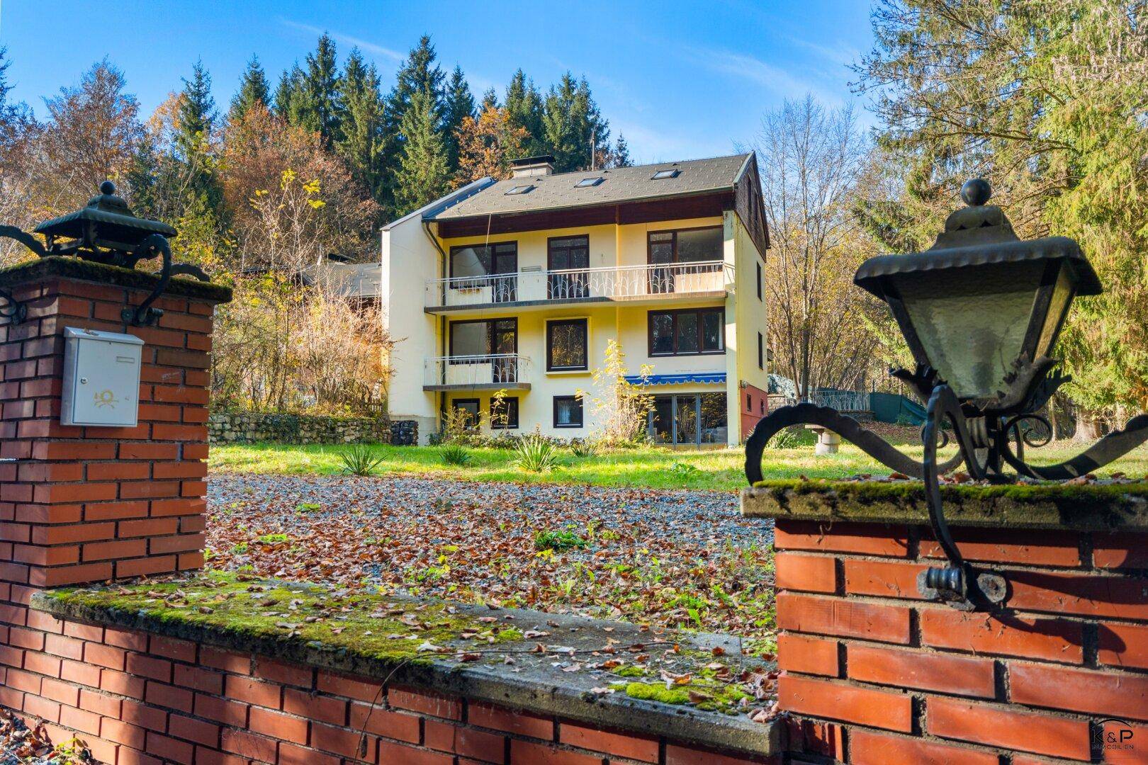 Ansicht des Gebäudes durch ein traditionelles Ziegelmauer-Tor mit Laternen in bewaldeter Umgebung.