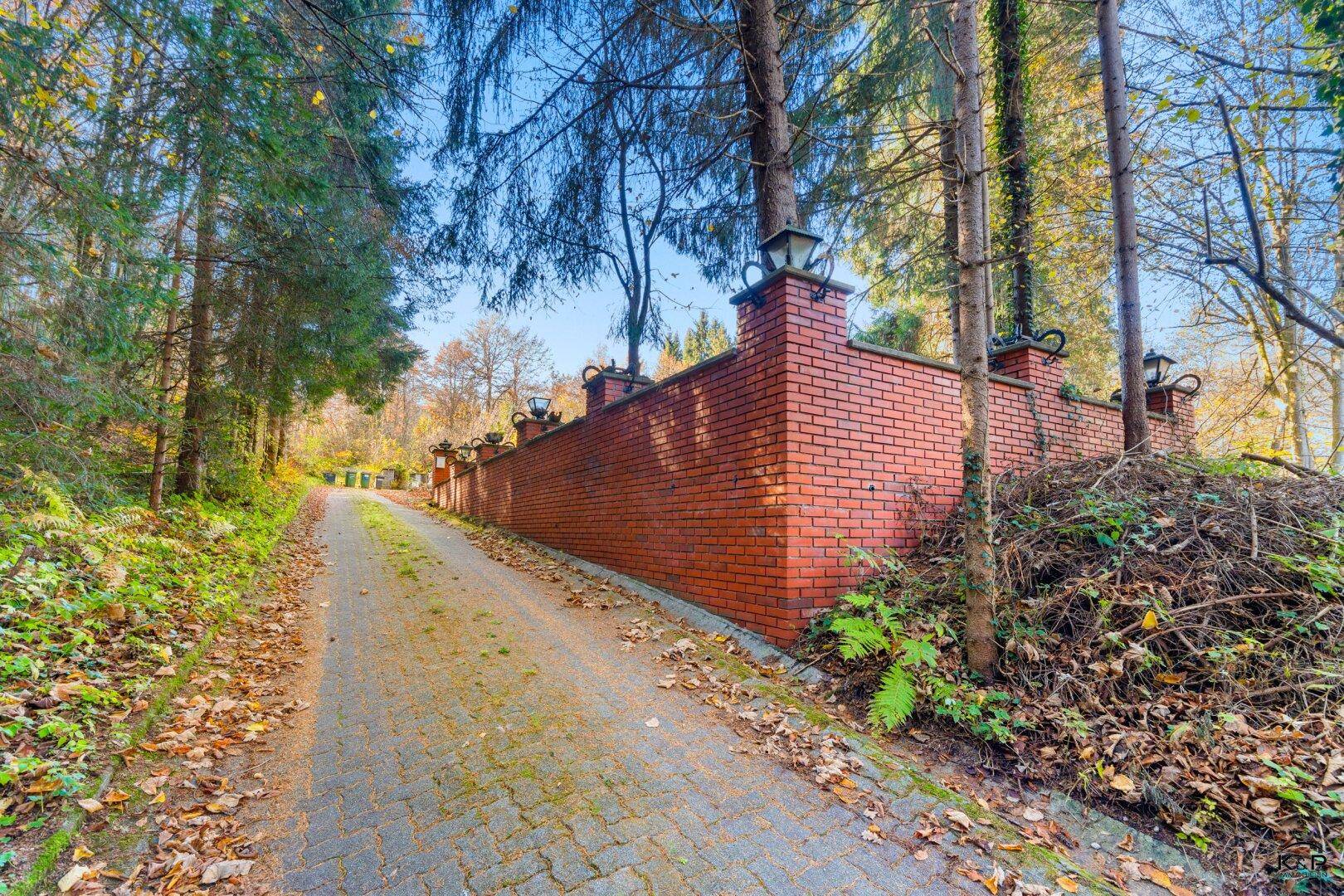 Zufahrt zum Grundstück mit einer langen Ziegelmauer und dekorativen Laternen, umgeben von Bäumen.
