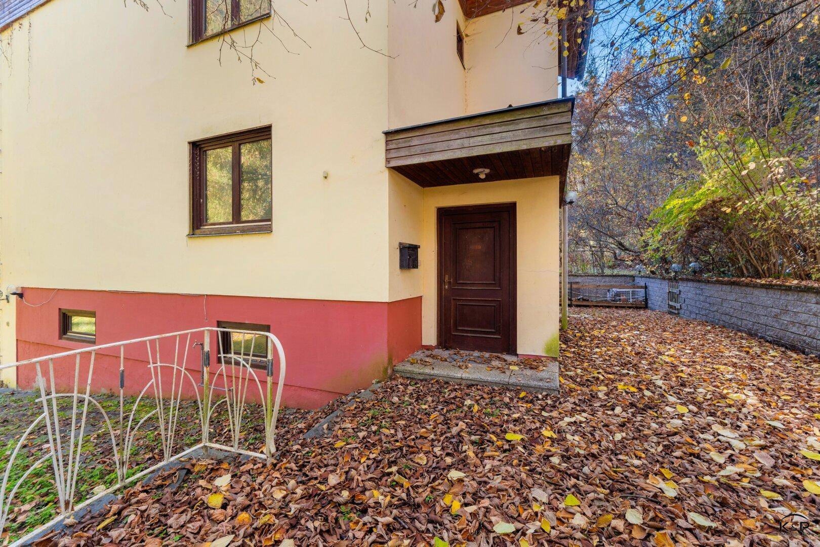 Haupteingang des Gebäudes mit Holztür und Treppenzugang, umgeben von Herbstlaub.