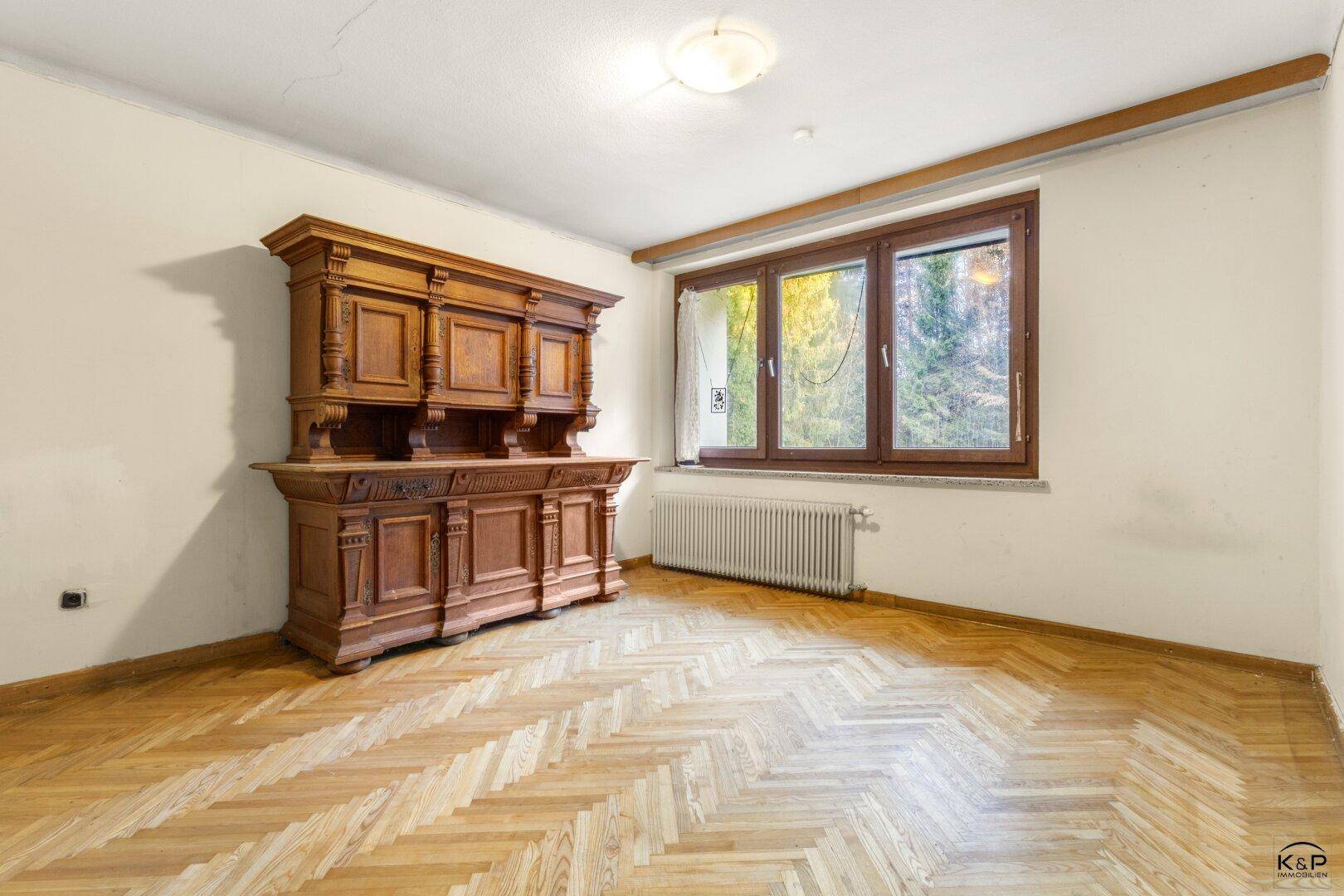 Geräumiger Raum mit Fischgrätparkett, einem großen Holzschrank und Blick ins Grüne.