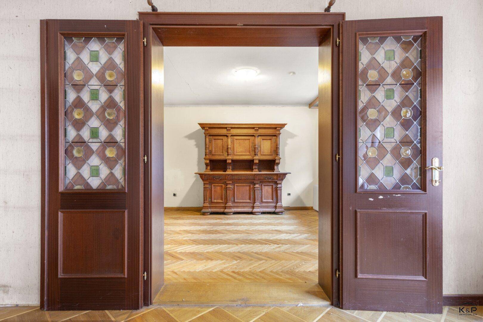 Antike Holztüren mit Buntglasfenstern öffnen sich zu einem Raum mit Fischgrätparkett und einem Holzschrank.