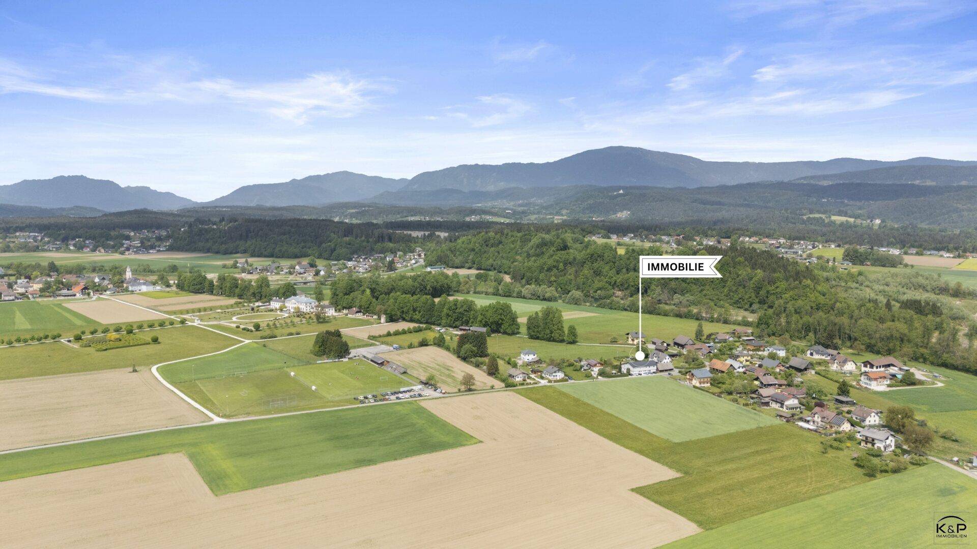 Weitläufige Luftaufnahme der Immobilie, eingebettet in eine grüne Landschaft mit umliegenden Bergen und Dorf.