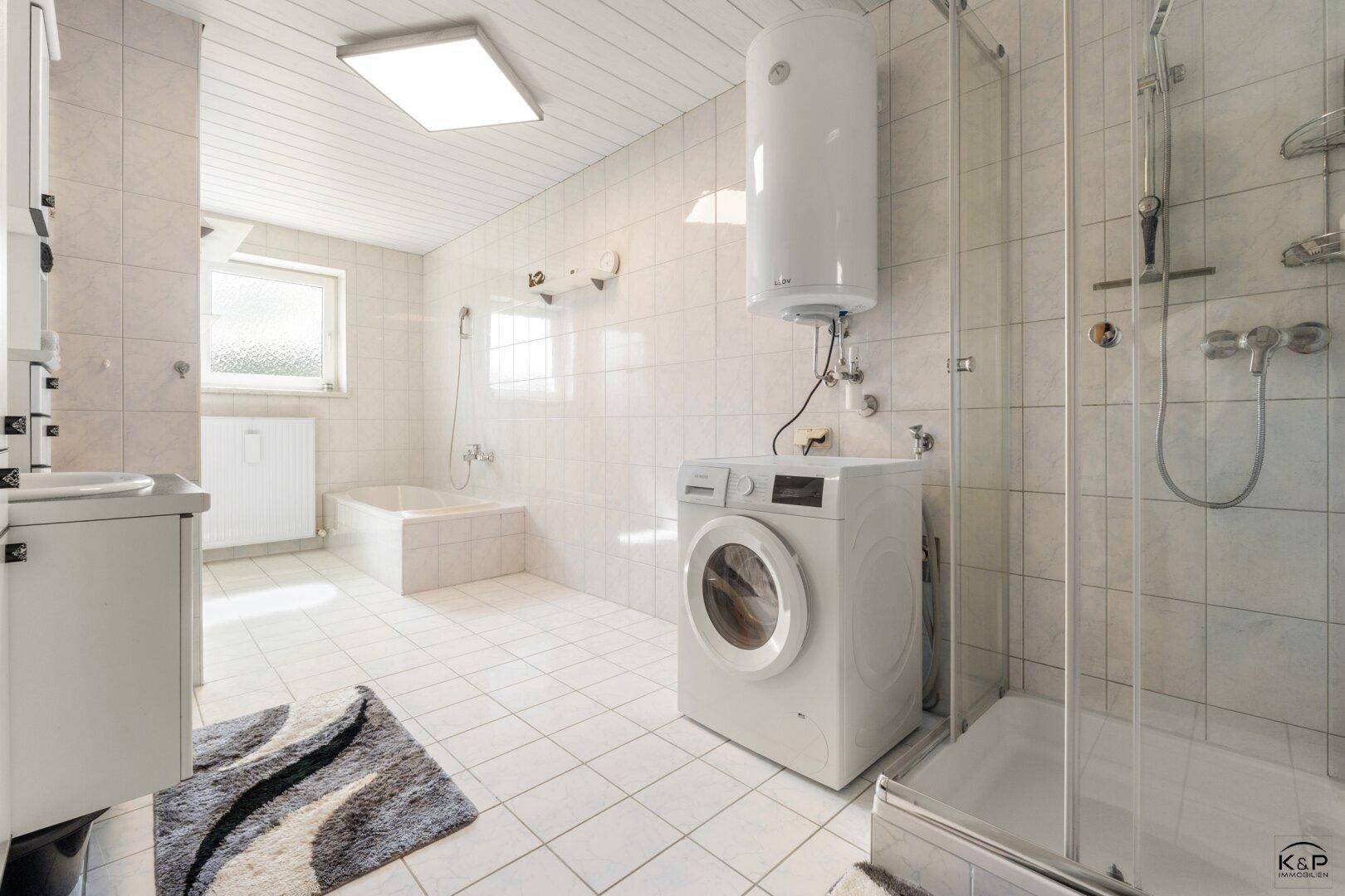 Helles Badezimmer mit weißen Fliesen, Badewanne, Dusche und Waschmaschine.