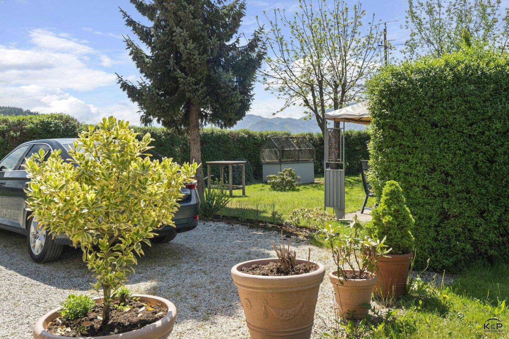 Kiesauffahrt mit Topfpflanzen und geparktem Auto, im Hintergrund der Garten und die Berge.