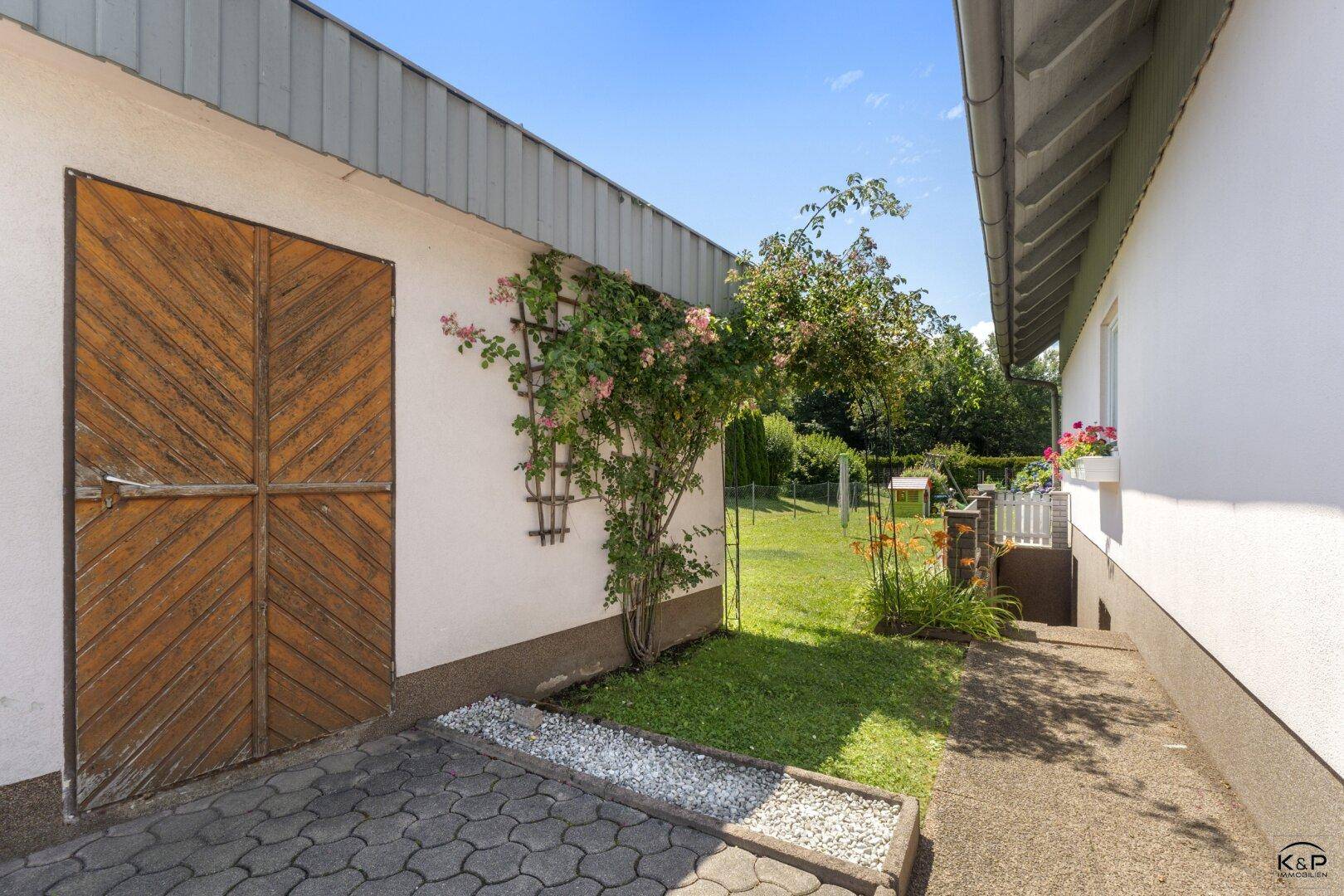Holzgaragentor neben einem grünen Gartenbereich mit blühenden Pflanzen und Kletterrosen.