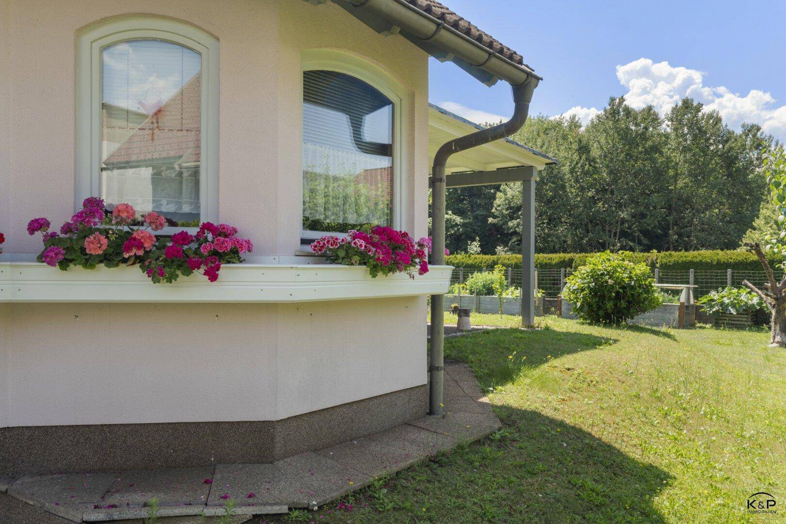 Hausfassade mit Blumenkästen an den Fenstern und Blick auf den grünen Gartenbereich.