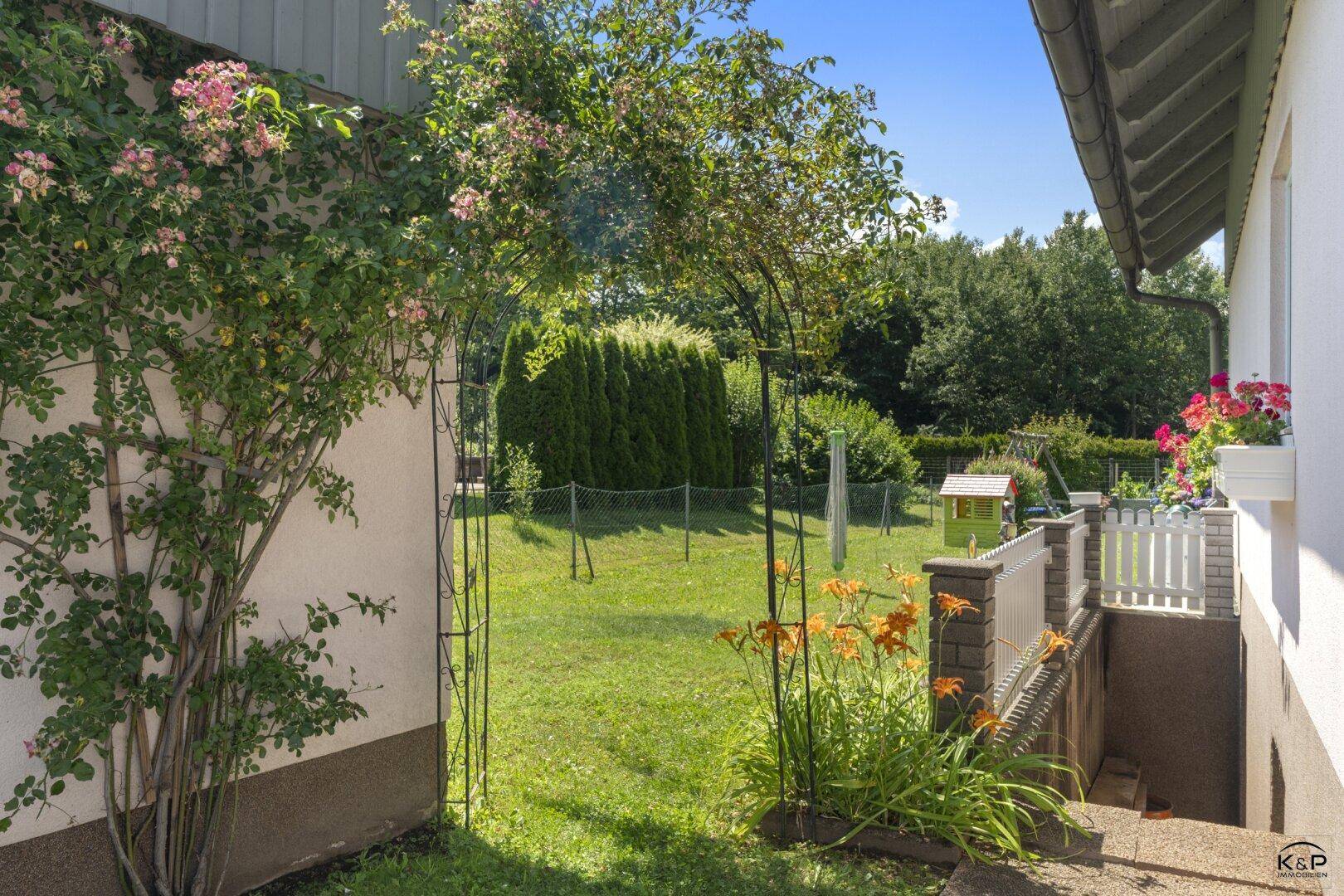 Seitlicher Gartenbereich mit Kletterrosen, Blumenbeeten und Zugang zum Kellerabgang.