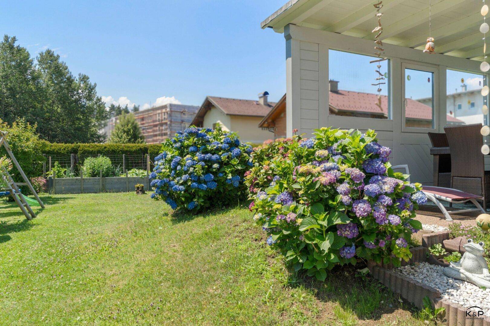 Großer, sonniger Garten mit blühenden Hortensien, Schaukel und einer überdachten Terrasse.