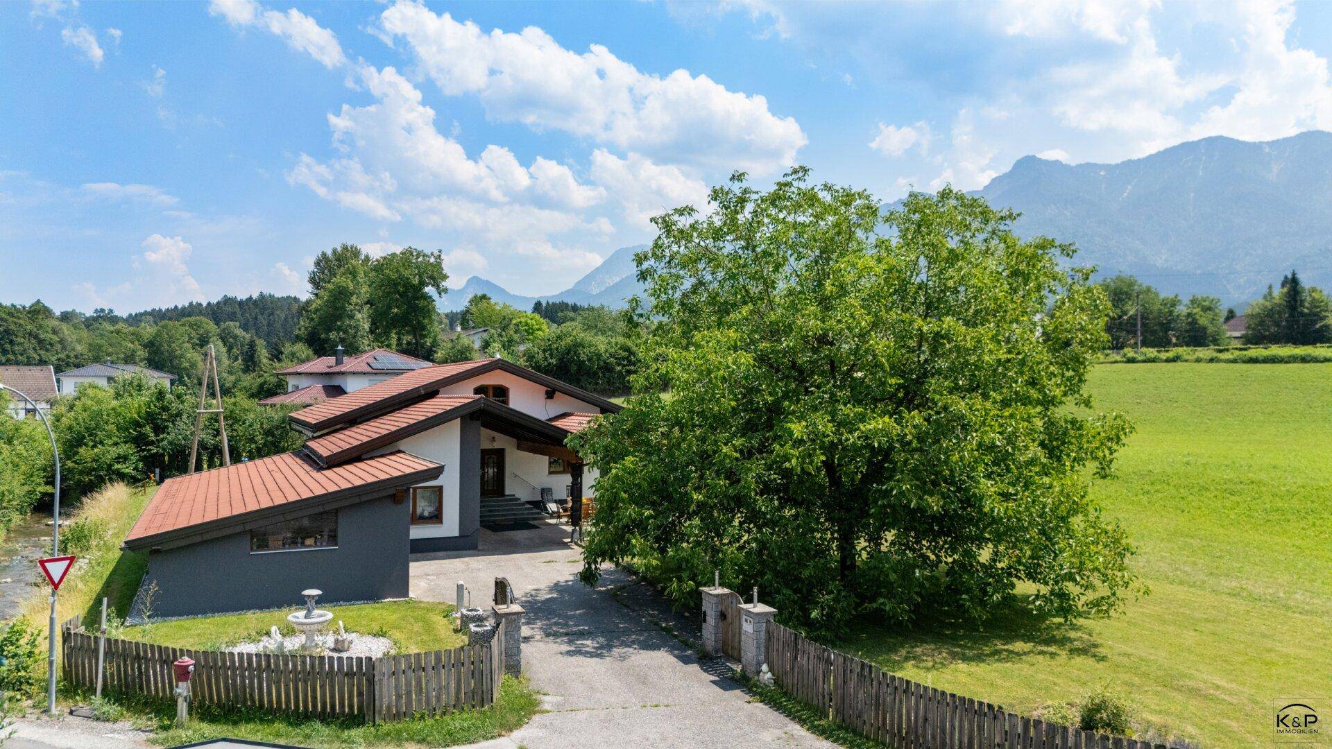 Einfahrt zum Haus mit großem Baum, umzäuntem Vorgarten und beeindruckendem Bergblick.