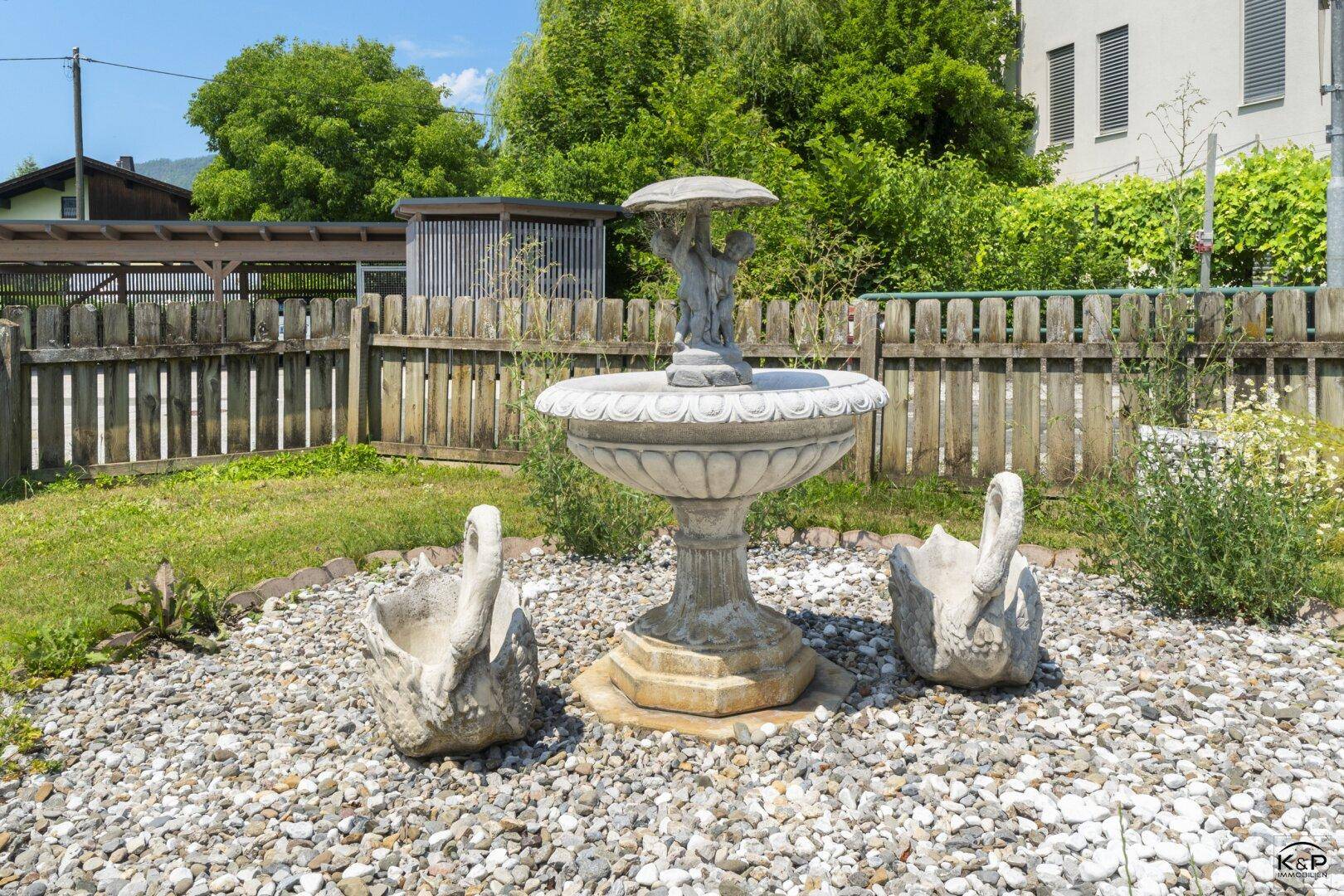 Dekorativer Gartenbrunnen mit Schwänen in einem Kiesbeet, umgeben von einem Holzzaun.